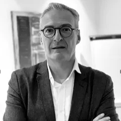 Black and white photo of a middle-aged man with glasses, wearing a blazer and white shirt, standing with arms crossed indoors.