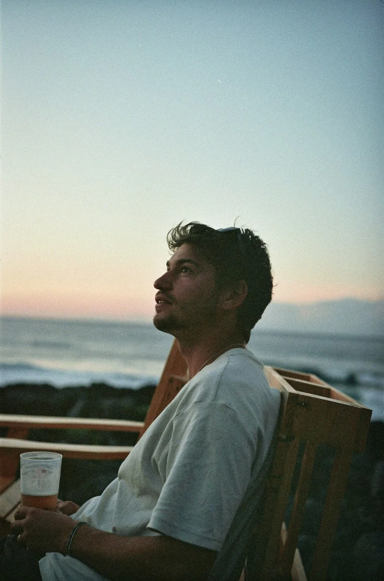 Rayane Zidane, freelance UX designer and Webflow developer, on a wooden bench by the sea at sunset, looking up